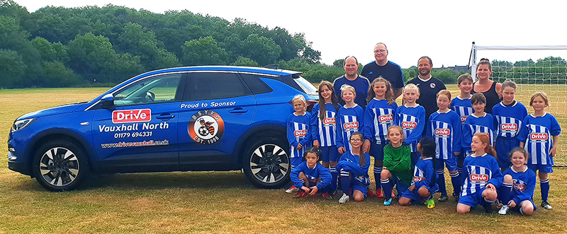 Photo of the BSYFC U9 Girls in their match kit sponsored by Drive Vauxhall Bristol North standing next to a Vauxhall Crossland X vehicle .