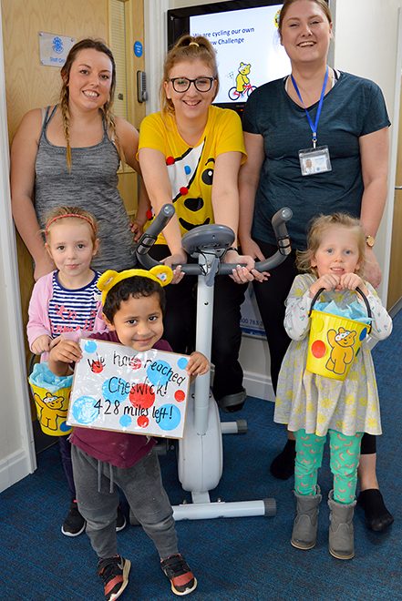 Photo of a member of staff on an exercise bike, surrounded by other members of staff and children.