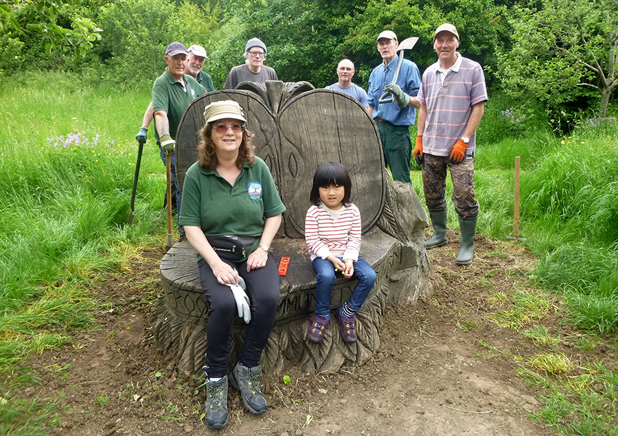 Volunteers from the Three Brooks Nature Conservation Group at the 'apple bench'. Photo of people standing around and sitting on a wooden sculpture.