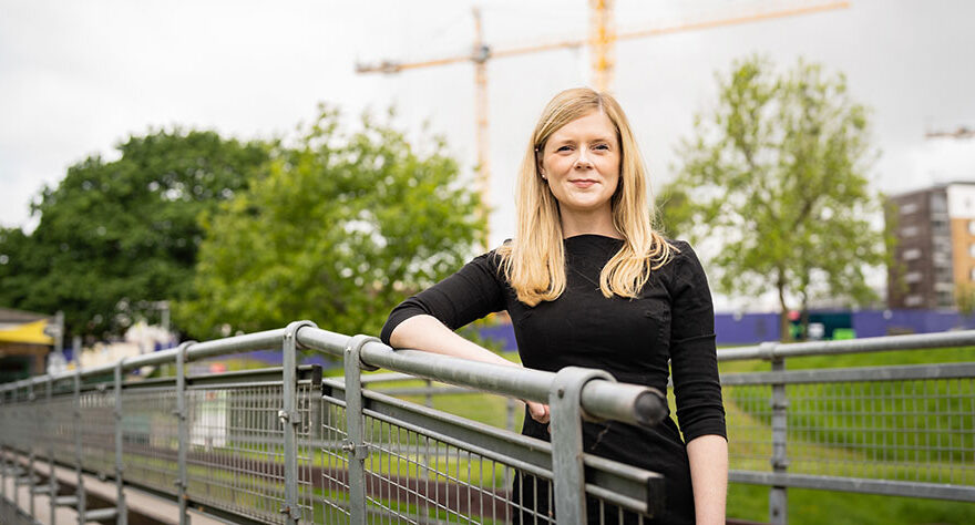 Photo of a woman in a black dress with one arm resting on a railing. Two construction cranes visible in the background.