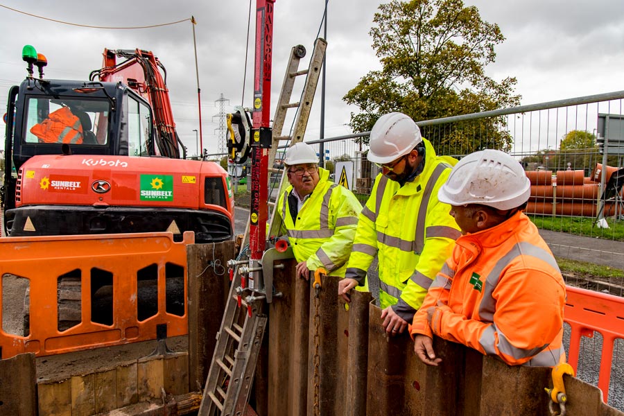 Cllr Steve Reade (left) and engineers discuss the scheme to replace 80 metres of damaged drainage pipe, 3 metres below the road surface. Photo of a group of people in hard hats at a road excavation site.