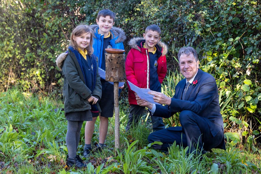 Photo of three school pupils and a man (kneeling) gathered around a bee-friendly object in a garden.