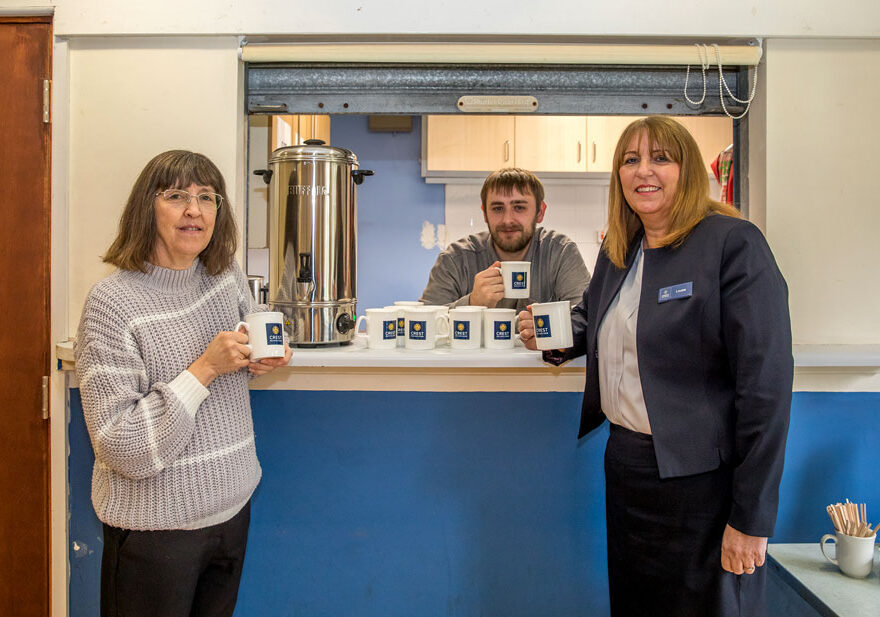 Photo of three people standing near a tea urn at a serving hatch.